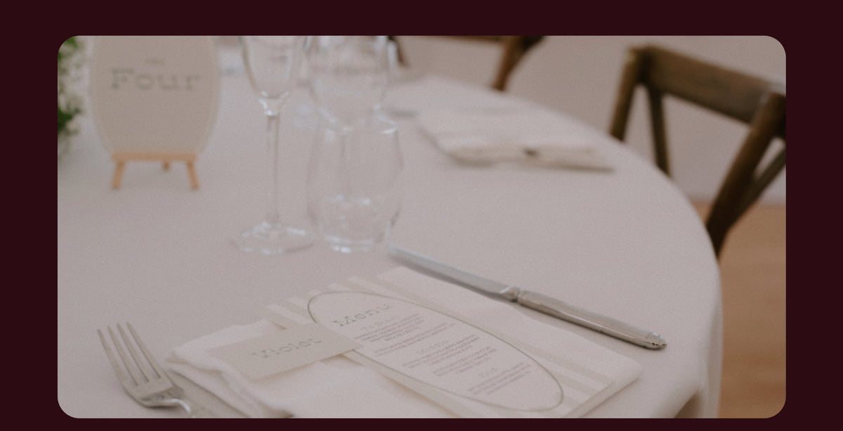 Wedding stationery on a white tablecloth with a printed menu, folded napkin, cutlery, glassware and wooden chairs