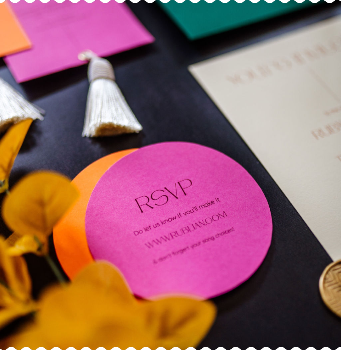 Round pink stationery card with black RSVP lettering, layered over an orange card, tassels and dried leaves
